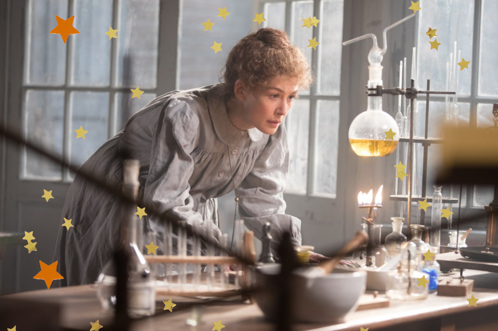 Foto de divulgação do filme Radioactive, protagonizado por Rosamund Pike; a atriz está caracterizada dentro de um laboratório de química, olhando para um vidro que consta uma substância laranja