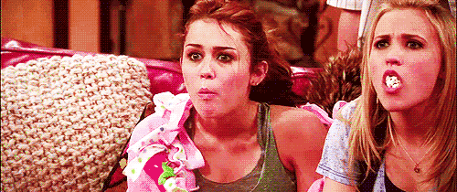 Gif de Miley Cyrus e Emily Osment como Miley Stewart e Lilly Truscott em Hannah Montana. Elas estão sentadas no sofá, cuspindo a pipoca que está na borra. Miley está usando uma regata verde e um cobertor rosa em cima do ombro esquerdo, Lilly está com um casaquinho branco. As duas têm expressão facial de surpresa.
