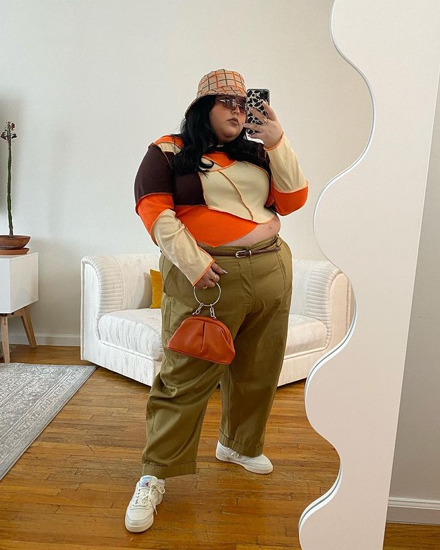 Garota usando blusa de manga longa de patchwork nas cores laranja, bege e marrom. Ela está usando uma calça baggy bege escura, com cinto fino marrom, tênis branco e um bucket hat xadrez laranja e marrom. Ela está em pé, tirando foto em frente a um espelho com o celular na mão, e uma bolsa pequena laranja na outra, além de óculos de sol com lentes marrons.