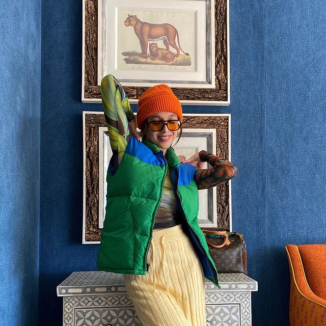 A influenciadora americana Emma Chamberlain usando colete verde e azul por cima de blusa segunda pele preta com tons de verde, calça de tricô amarela, gorro laranja e óculos de sol com lentes alaranjadas. Na foto, do joelho para cima, ela está com os dois braços dobrados para trás.