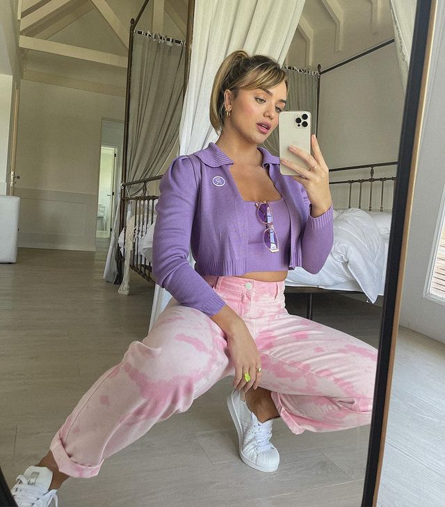 Rafa Kalimann está usando top e casaquinho no mesmo tom de lilás, uma calça rosa tie-dye e um tênis branco. Ela está agachada em frente ao espelho com o celular próximo ao rosto.