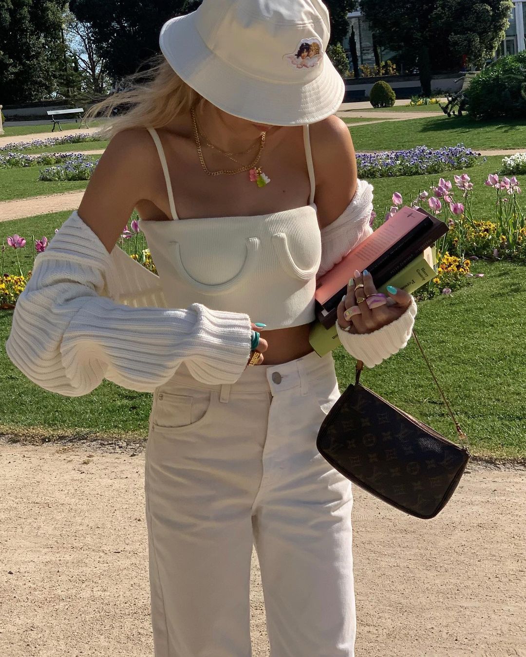 Garota usando top cropped off white com calça, cardigan e chapéu da mesma cor. O chapéu cobre seu rosto enquanto ela olha para baixo. Uma das mãos está segurando livros e uma bolsa preta.