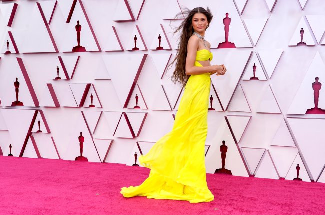 Zendaya usando vestido amarelo esvoaçante no Oscar 2021. Ela está com o corpo de lado, cabelo solto aos ventos, as mãos na frente do corpo e olhando para o lado com expressão facial séria.