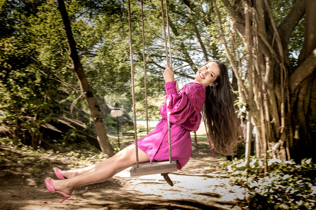 Juliette, vencedora do BBB21, em um balanço no meio de uma floresta usando um vestido rosa, sorrindo para câmera