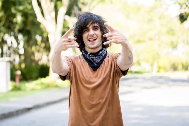 Fiuk posando para foto com as mãos fazendo símbolo do rock usando camiseta marrom e bandana preta amarrada no pescoço