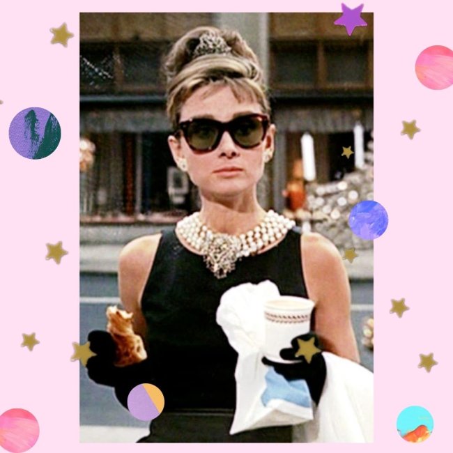 Audrey Hepburn como Holly, no filme Bonequinha de Luxo, segurando um saco branco com um copo de café e croissant, usando o vestido preto com colares, e óculos preto, com expressão séria.