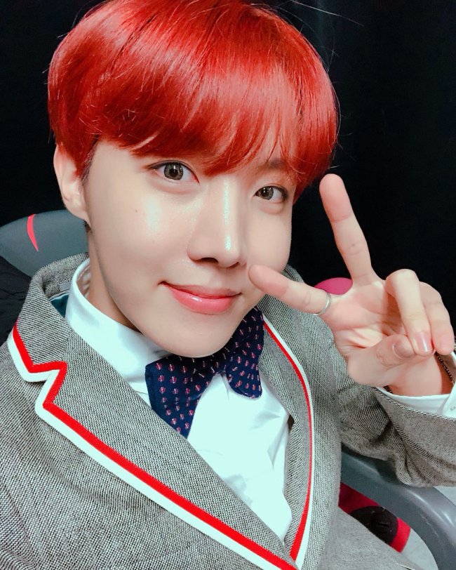 J-hope, do grupo BTS, com cabelo colorido, fazendo sinal de paz e amor, usando gravata borboleta e blazer cinza com lista vermelha e branca.