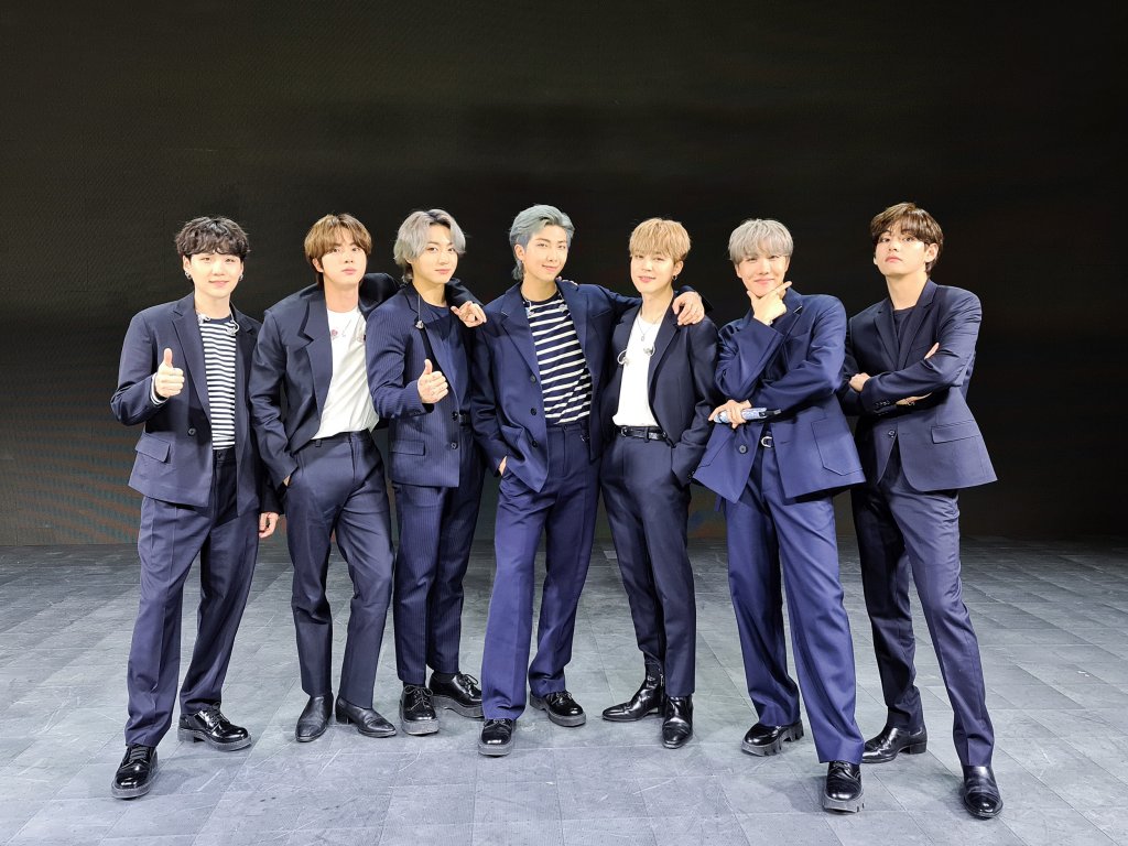 Os sete integrantes do BTS alinhados na horizontal em imagem promocional do single Butter; eles usam ternos em tons de azul escuro em fundo preto com chão cinza