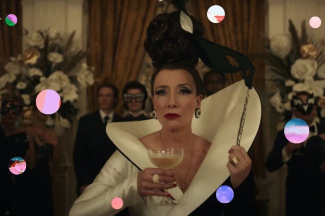 Emma Thompson como Baronesa no filme Cruella, com roupa branca e preta, segurando uma mascara preta e uma taça na outra mão. Sua expressão é séria.