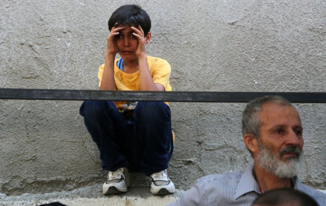 Garotinho palestino ajoelhado no chão, com as mãos nas laterais dos olhos, com cara te desespero