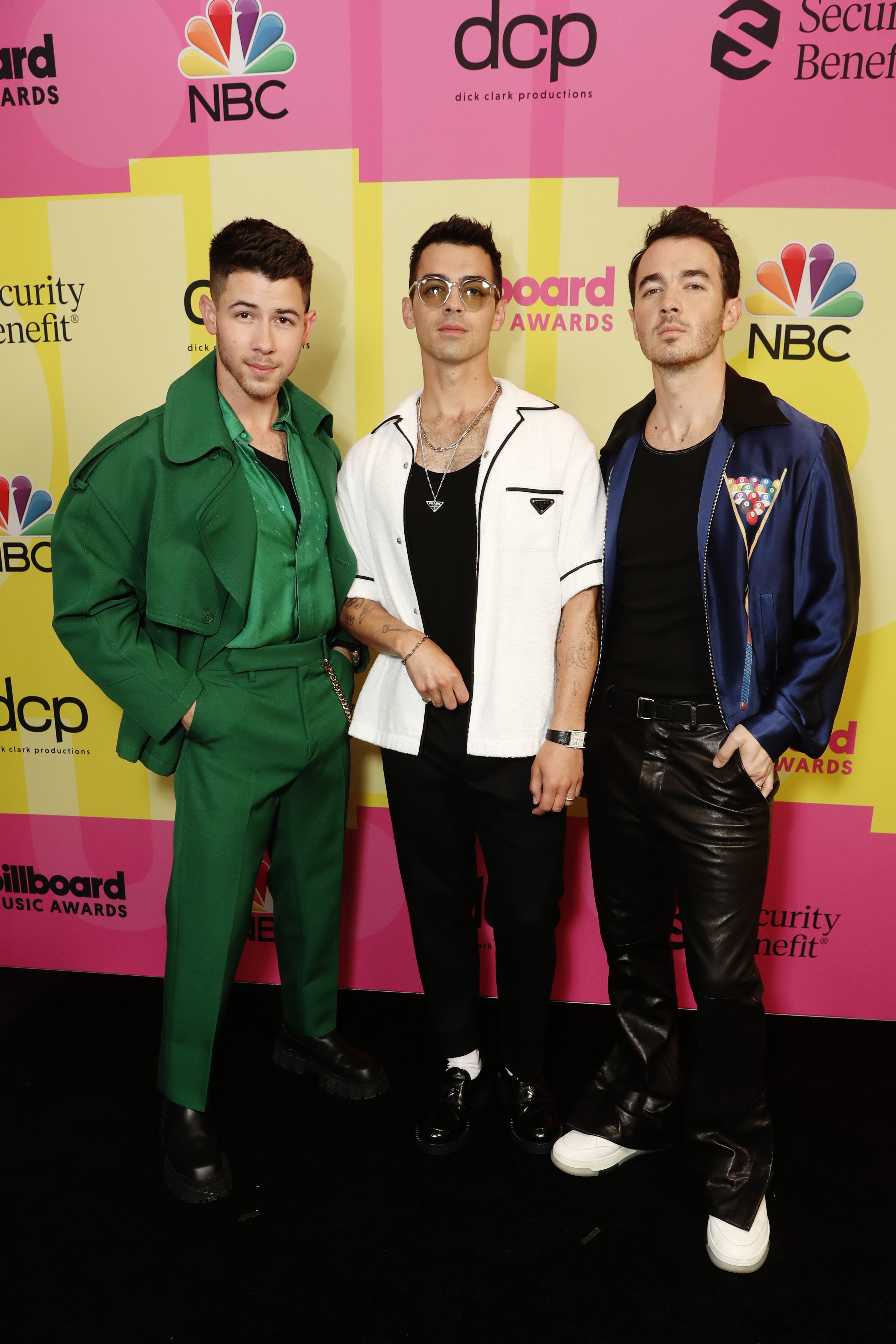 Os Jonas Brothers no Billboard Music Awards 2021. Nick Jonas está com um look todo verde bandeira, Joe Jonas está de preto com uma camisa branca por cima, e Kevin Jonas está de preto com uma jaqueta azul-marinho.