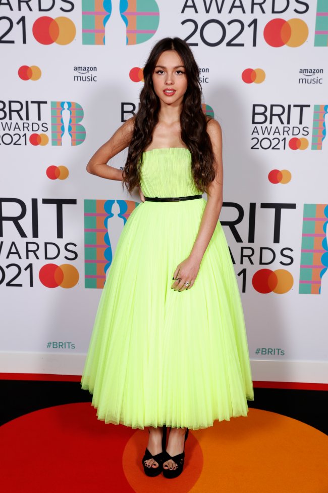 Olivia Rodrigo no tapete vermelho do BRIT Awards 2021. Ela está usando um vestido em tom de verde-limão sem alças e com saia rodada e um cinto preto, além de sandália preta de salto. Seu cabelo está solto, expressão facial séria, uma das mãos na frente do corpo, e a outra na cintura.