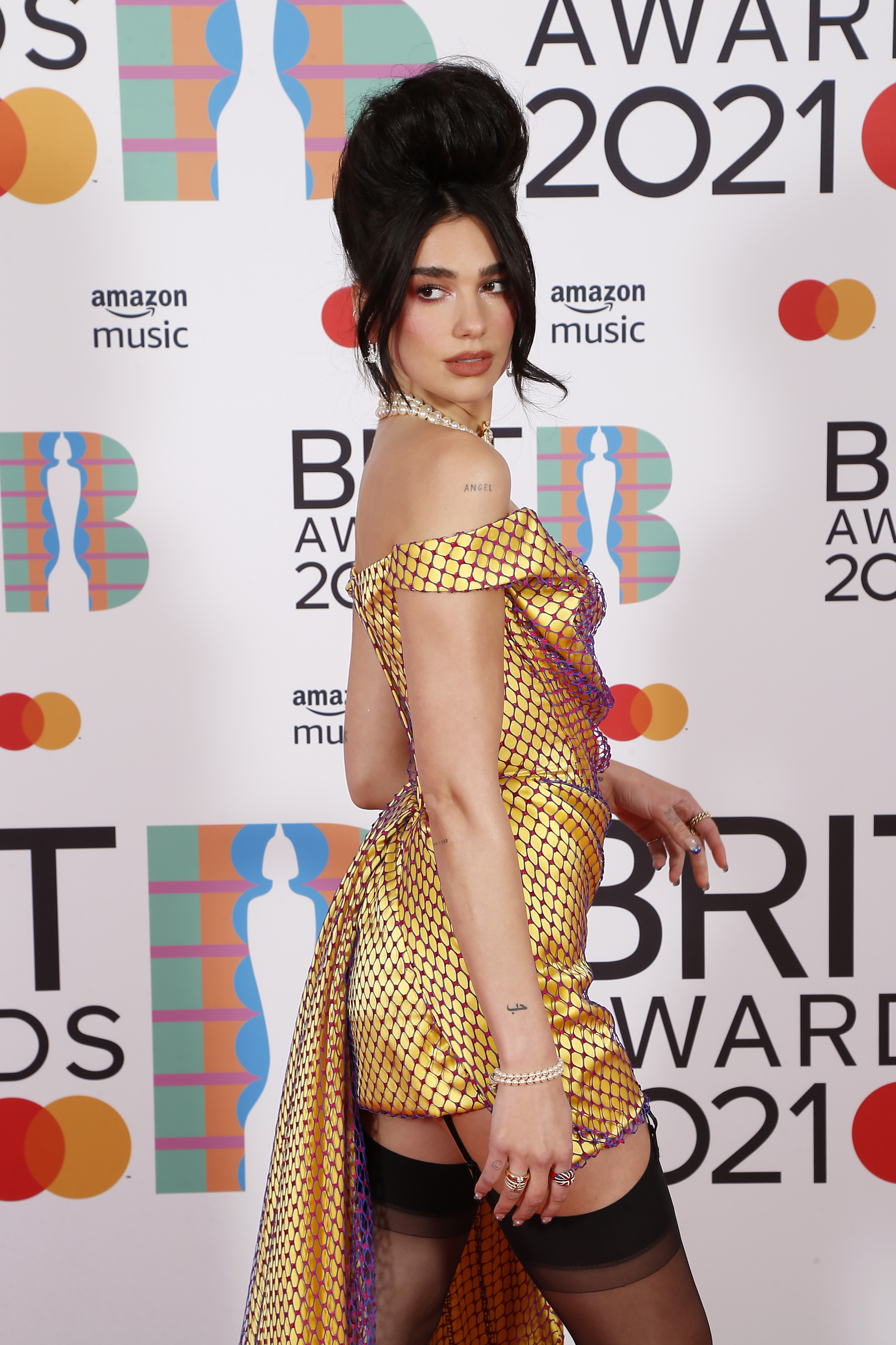 Dua Lipa no BRIT Awards 2021. Ela está usando um vestido dourado curto com uma cauda lateral e uma renda de furinhos roxos por cima. Na foto, ela está virada para um dos lados, olhando por cima de um dos ombros. Seu cabelo está preso em um coque volumoso.