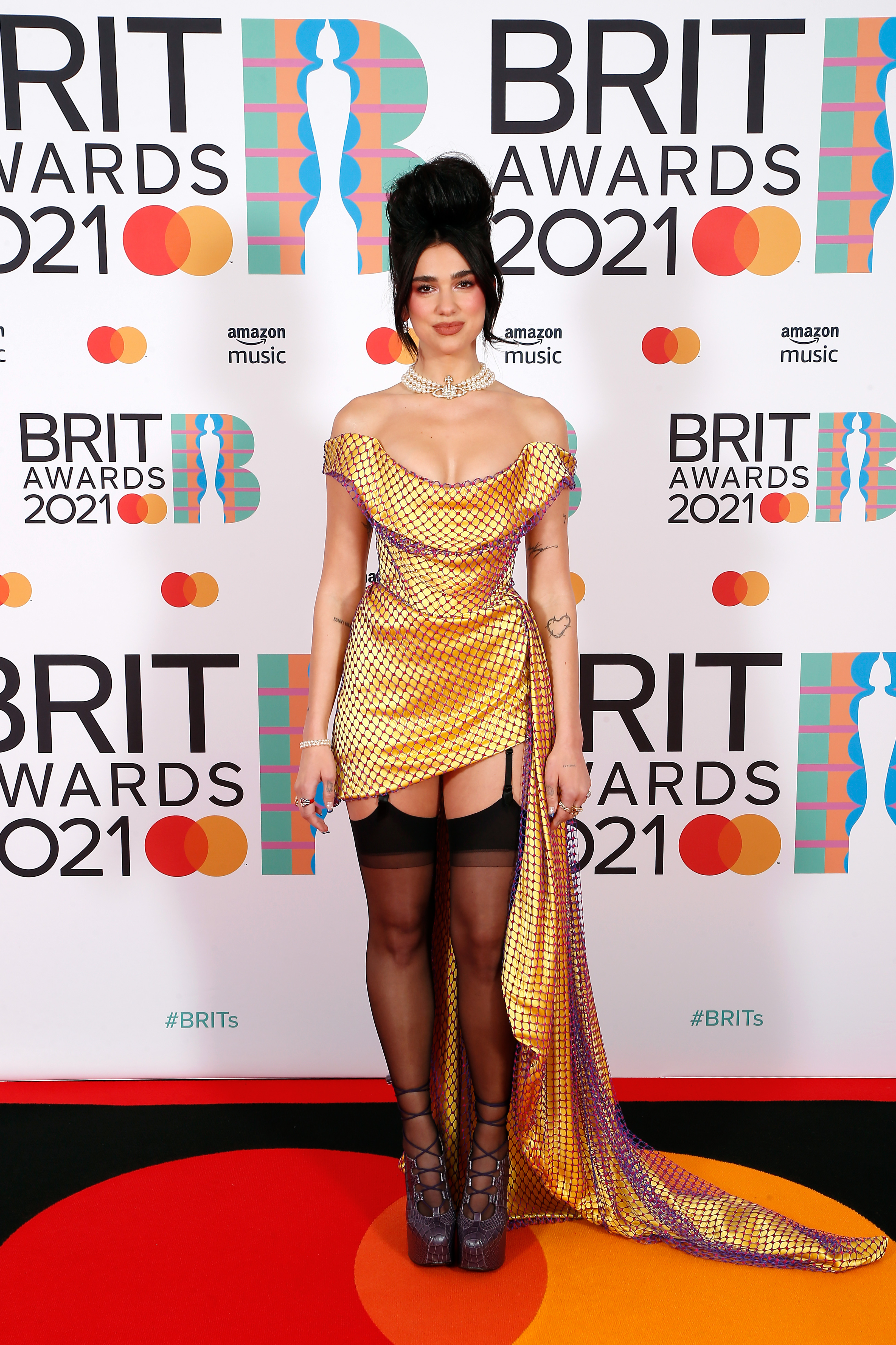 dua-lipa-brit-awards-2021 Dua Lipa no tapete vermelho do BRIT Awards 2021. Ela está com um leve sorriso sem mostrar os dentes, usando um vestido dourado com uma rede de furinhos roxos por cima, uma meia-calça preta e um sapato de salto. Também está com colar de pérolas e cabelo preso em um coque alto bem volumoso.