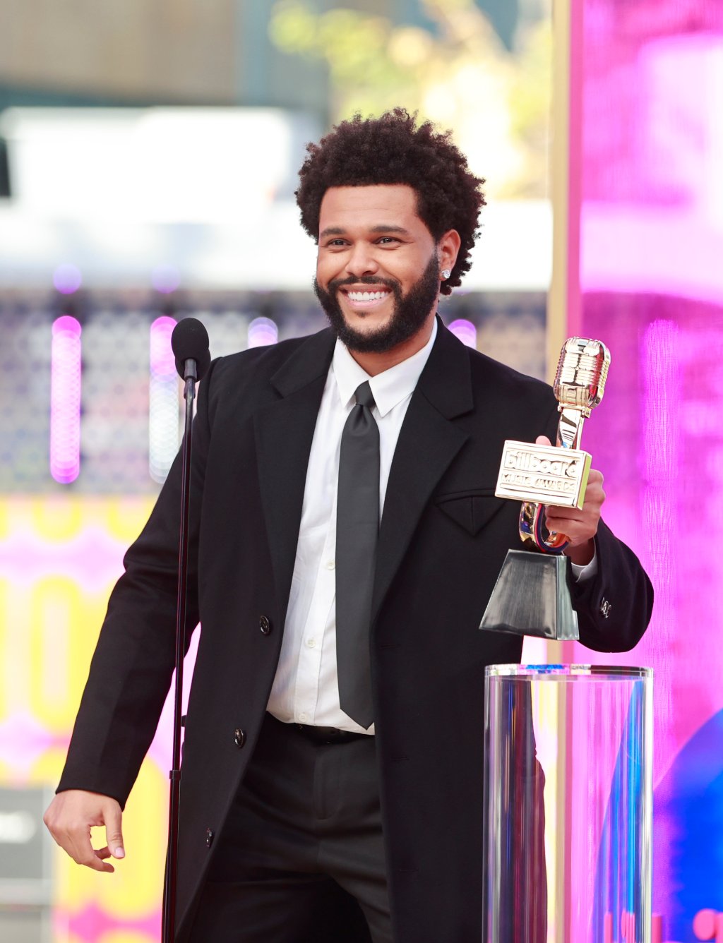 The Weeknd segurando prêmio no Billboard Music Awards 2021. Ele está sorrindo, usando camisa branca, gravata preta, sobretudo preto e calça preta, e há um microfone em sua frente.