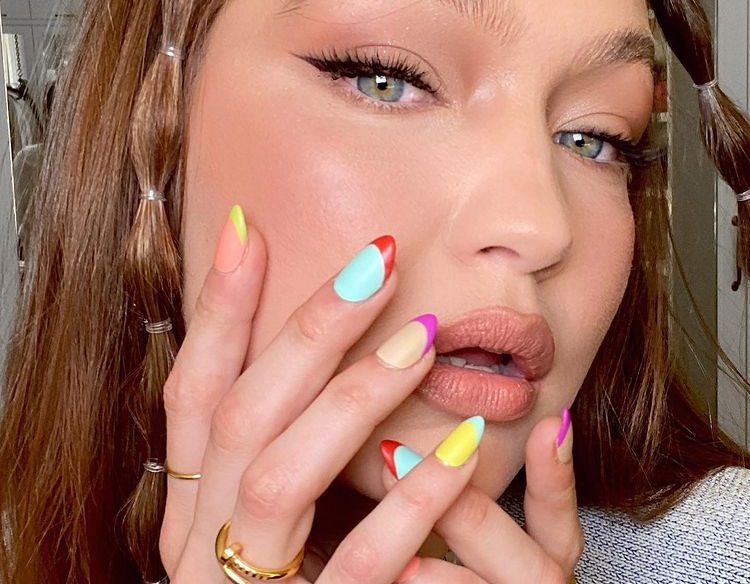 Gigi Hadid com nail art de francesinha colorida, posando com as mãos no rosto para evidenciar as unhas, suas unhas estão com esmalte azul, amarelo, laranja, roxo e rosa. Sua expressão é seria. Nos cabelos ela tem duas mechas com bubble hair.