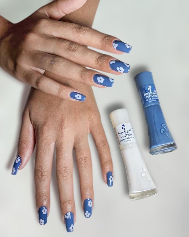 Duas mãos ao lado de um esmalte branco e outro azul. Nas unhas com florzinha branca e fundo azul.