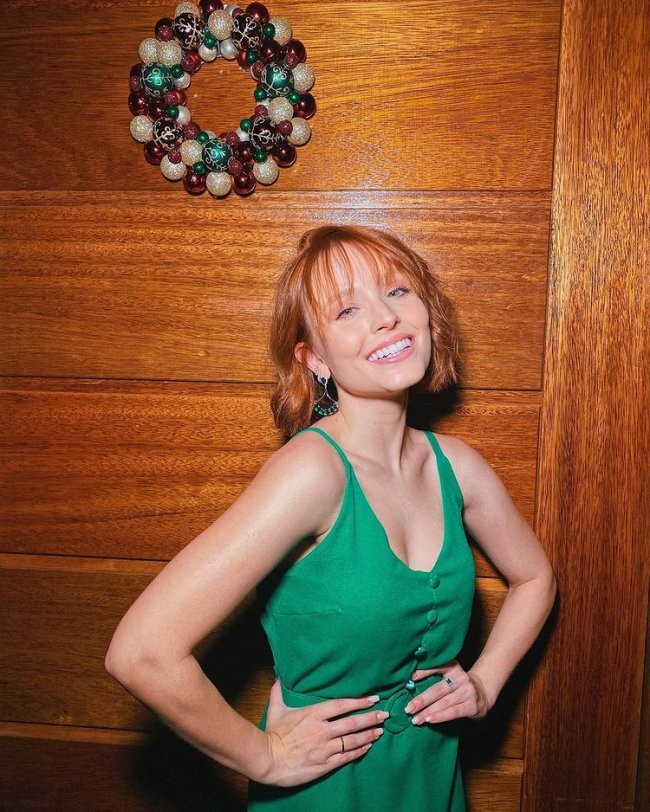 Larissa Manoela posando com as duas mãos na cintura e sorridente, usando blusa verde e em frente a porta marrom com decoração natalina.