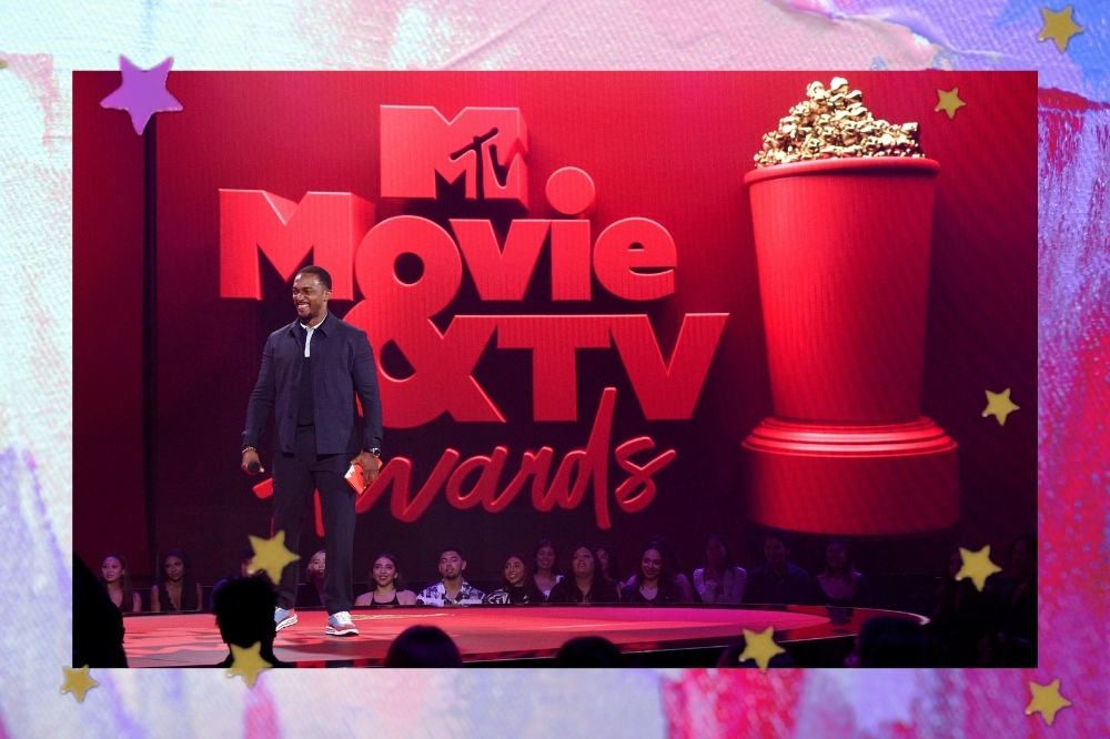 Imagem com a fachada da premiação Mtv Movie & Tv Awards 2021 que é toda vermelha, com o nome da premiação em relevo e um grande balde de pipoca ao lado, o ator Anthony Mackie aparece ao lado sorridente.