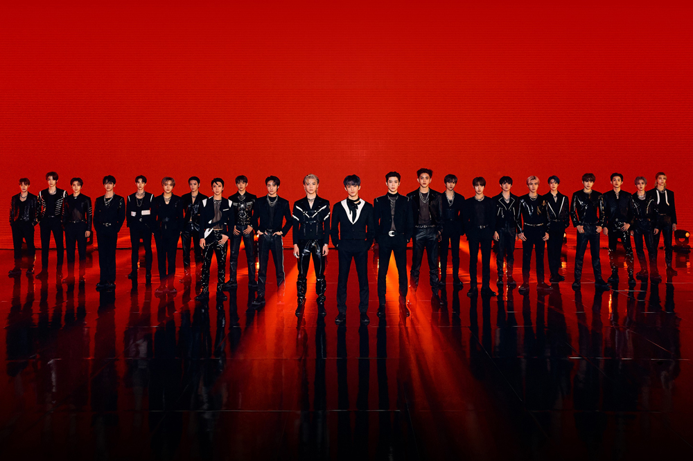 Supergrupo NCT em um fundo vermelho, os integrantes estão alinhados na diagonal formando uma espécie de triângulo com um integrante no meio; todos vestem roupas completamente pretas, algumas com detalhes brancos