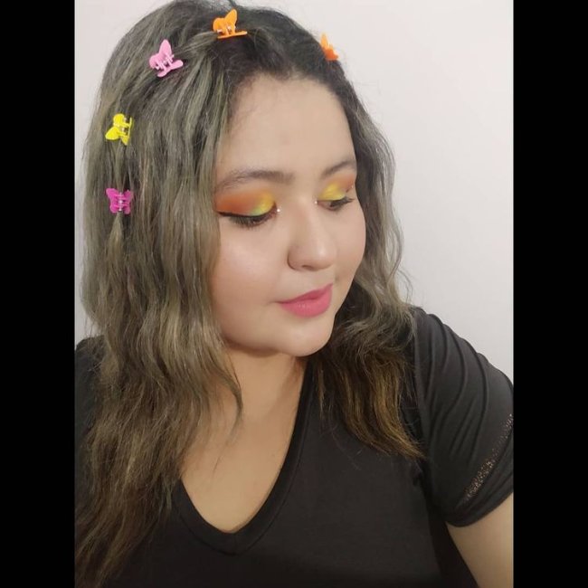 Penteado com borboleta colorida Jovem olhando para baixo com sombra amarela e laranja, usando presilha de borboleta na lateral do cabelo usando camiseta preta com uma expressão sorridente.