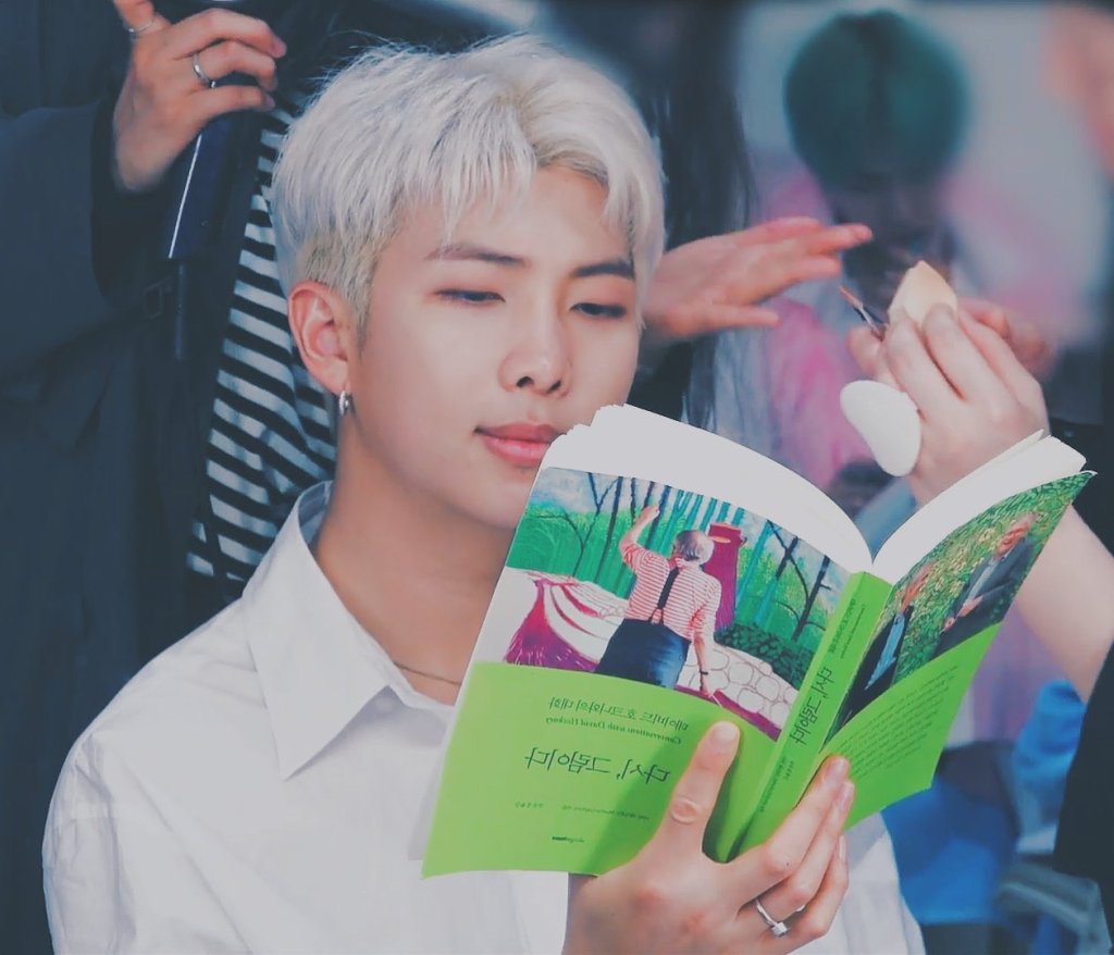 RMT lendo livro; ele usa uma camisa branca e seu cabelo está platinado, o cantor sorri levemente