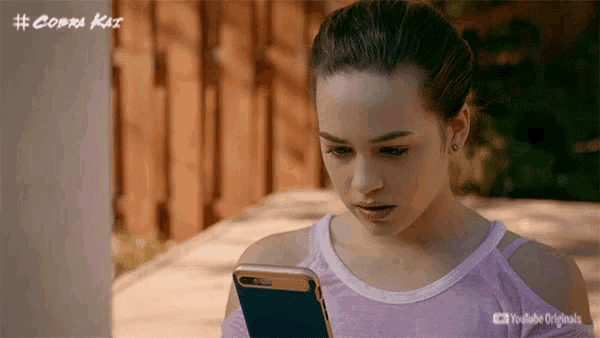 Garota olhando com cara de angustiada para a tela do celular; ela está com o cabelo preso em um rabo e usa camiseta branca