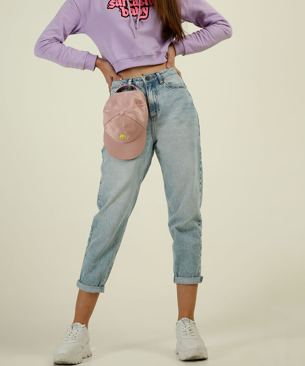 Garota usando calça mom jeans da coleção da CAPRICHO com a MARISA. Tem um boné rosa pendurado em um dos passadores da calça, ela está de tênis branco e dá para ver um pedaço do moletom lilás. Suas duas mãos estão na cintura.