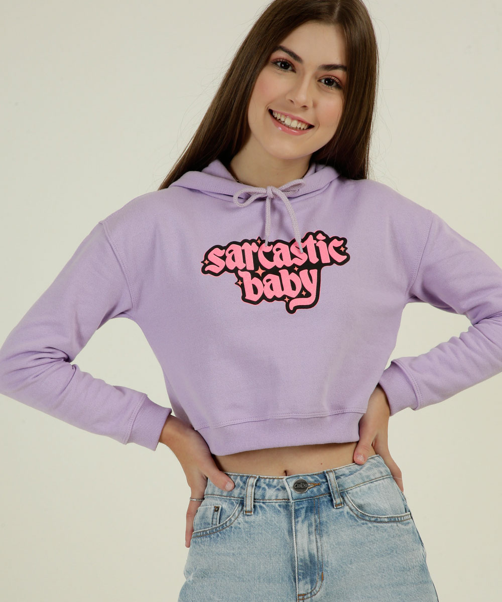 Garota usando blusa de moletom lilás escrito sarcastic baby da coleção da CAPRICHO com a MARISA. Ela está sorrindo, com as duas mãos apoiadas na cintura e também dá para ver um pedaço da calça jeans.