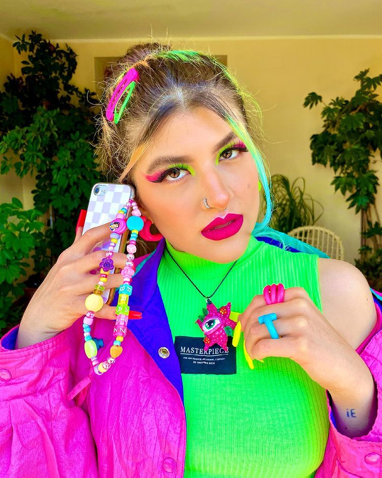 cordinha-celular-0ceanzs Garota usando top cropped verde neon e jaqueta roxa e azul. Ela está segurando seu celular, que tem uma cordinha de miçangas pendurada, perto da orelha, e sua expressão facial é séria. Sua maquiagem é verde neon e rosa, seu cabelo está preso e há presilhas rosa e verde neon na cabeça.