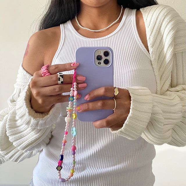 cordinha-celular-batninja Garota usando regata branca, cardigan off white e tirando foto na frente do espelho segurando o celular que tem uma cordinha de miçangas pendurada. Na foto, só dá para ver da cintura até a altura do pescoço.