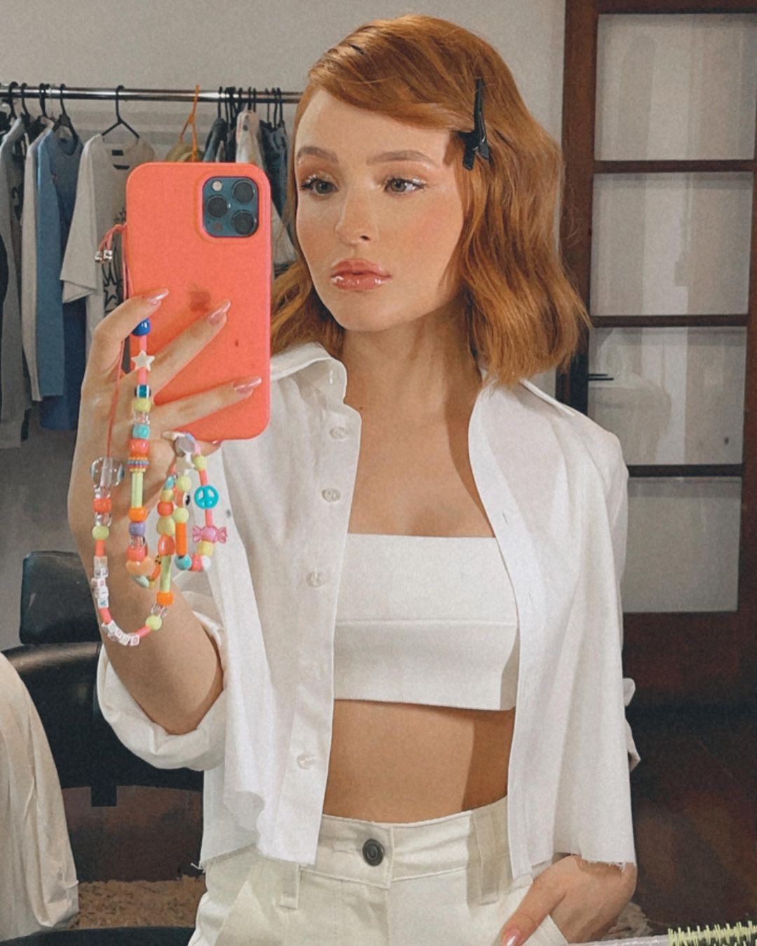Larissa Manoela usando conjuntinho todo branco formado por top cropped, calça e camisa. Ela está em frente ao espelho, segurando o celular que tem uma cordinha de miçangas pendurada, e com expressão facial séria.