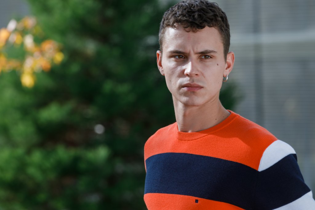 Ander em imagem promocional de Elite: Histórias Breves; ele usa uma camisa com listras laranjas e azul escuro e está com uma expressão fechada e série olhando para alguém ou algo próximo a ele