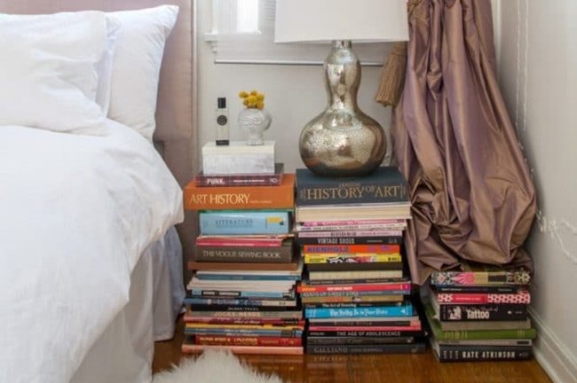 Quarto branco com uma mesa de cabeceira feita com pilhas de livros coloridos
