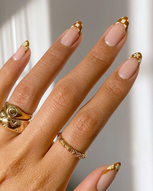 francesinha_xadrez Foto de uma mão com anéis dourados no dedo anelar e indicador. A unha está com uma nail art xadrez com francesinha na cor caramelo e branco.