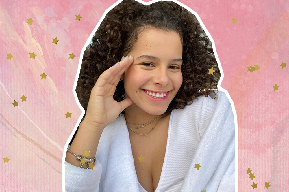 Colagem com foto de Gabriella Saraivah posando com a mão no rosto e sorrindo para câmera; ela usa uma roupa branca e está com a parte de cima do cabelo presa; o fundo é uma textura em tons de rosa e branco com estrelas amarelas de decoração