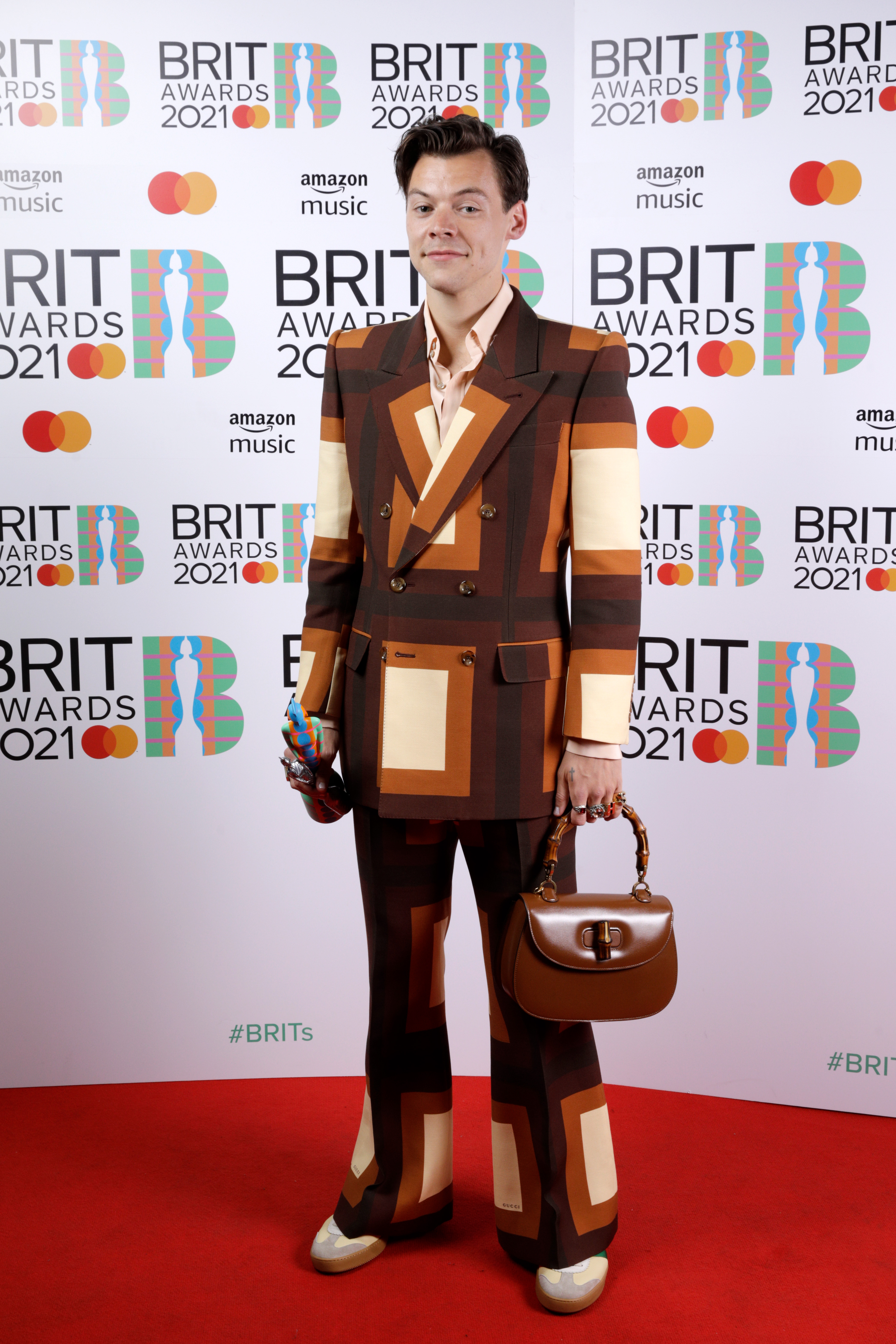 Harry Styles no tapete vermelho do BRIT Awards 2021. Ele está usando um terninho com xadrez em tons de marrom e bege. Com uma das mãos, ele segura uma bolsa marrom de alça curta, e com a outra uma estatueta da premiação. Ele está sorrindo levemente olhando para a câmera.Harry Styles no tapete vermelho do BRIT Awards 2021. Ele está usando um terninho com xadrez em tons de marrom e bege. Com uma das mãos, ele segura uma bolsa marrom de alça curta, e com a outra uma estatueta da premiação. Ele está sorrindo levemente olhando para a câmera.