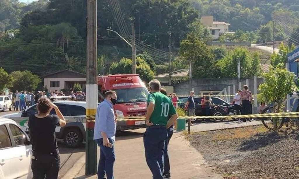A imagem mostra um homem de camisa azul conversando com outro homem de camisa verde. Ao fundo, está uma ambulância na porta de uma escola e um grupo de pessoas