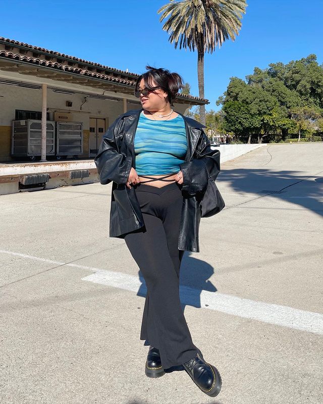 Foto de uma mulher na rua. Ela usa uma jaqueta de couro preta, blusa azul, calça legging bailarina, coturno e um óculos de sol vintage. Ela está com as mãos na cintura e olha para o lado.