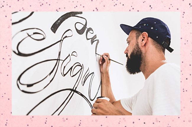 Um homem barbudo e de boné desenha algumas letras cursivas, conhecidas como lettering, numa parede branca