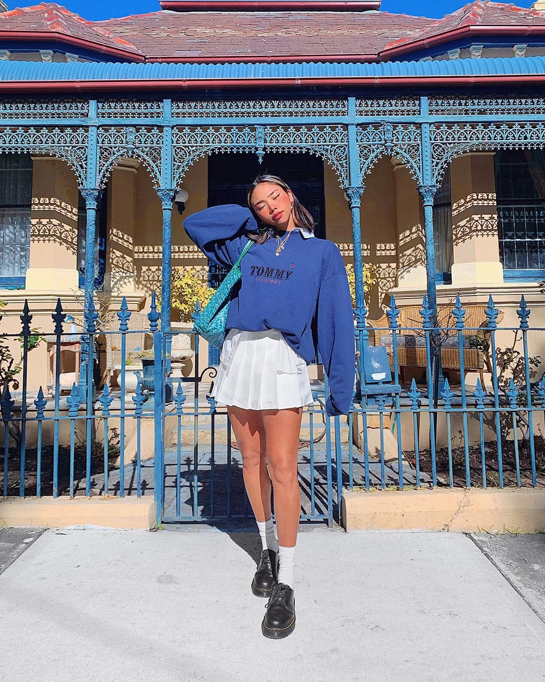 Foto de uma mulher em frente a uma casa com o portão azul. Ela usa um moletom vintage azul, saia colegial branca, sapato preto com meia branca. Ela está de olho fechado e cabeça apoiada no braço direito.