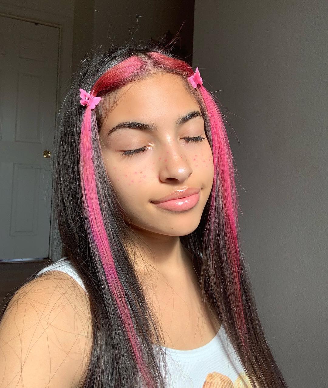 mecha-frontal-colorida-rosa Garota sorrindo com os olhos fechados para foto. Ela está vestindo uma regata branca, gloss nos lábios e cabelo preto com mechas frontais cor-de-rosa presas em dois acessórios de borboletas.
