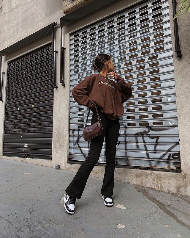 Foto de uma mulher na calçada. Ela veste um moletom vintage marrom, calça bailarina preta e tênis preto e branco. Ela olha para o lado e faz pose.