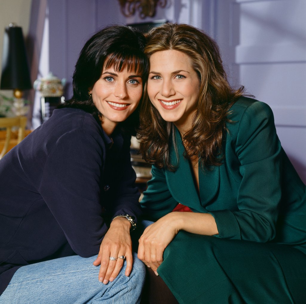 Courteney Cox, à esquerda, como Monica Geller e Jennifer Aniston, à direita, como Rachel Green em Friends. Courteney está usando um suéter azul e uma calça jeans e está sorrindo. Jennifer está usando um conjuntinho verde esmeralda de alfaiataria e sorrindo. Elas estão com as cabeças encostadas uma na outra.