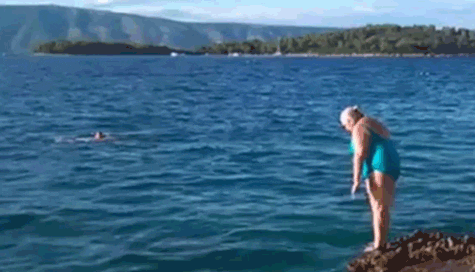 Idosa vestindo um maiô azul turquesa mergulha de cabeça no mar, mas acaba dando uma barrigada na água