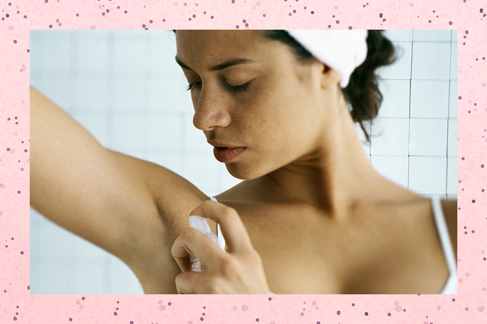 No banheiro, mulher usa faixa branca no cabelo enquanto passa desodorante em uma das axilas. Ela está olhando para baixo, com expressão facial séria, em direção à axila. O fundo da montagem é rosa com bolinhas.