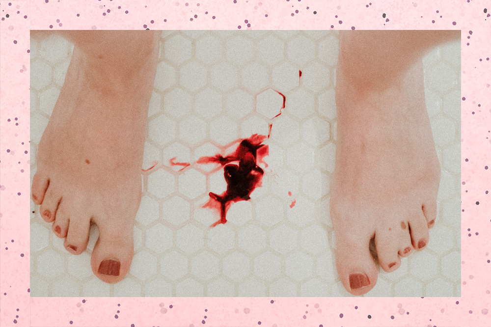 Na imagem, aparecem dóis pés com unhas pintadas de vermelho. Entre eles, uma mancha de sangue menstrual no chão branco.