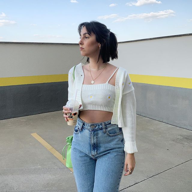 twin-set-cardigan-isabelabaccarini Garota usando top branco com cardigan branco com algumas florzinhas aplicadas e uma calça jeans de cintura alta e lavagem clara. Ela está olhando para o lado, com uma bolsa verde pendurada em um dos ombros e segurando um copo com uma mão.