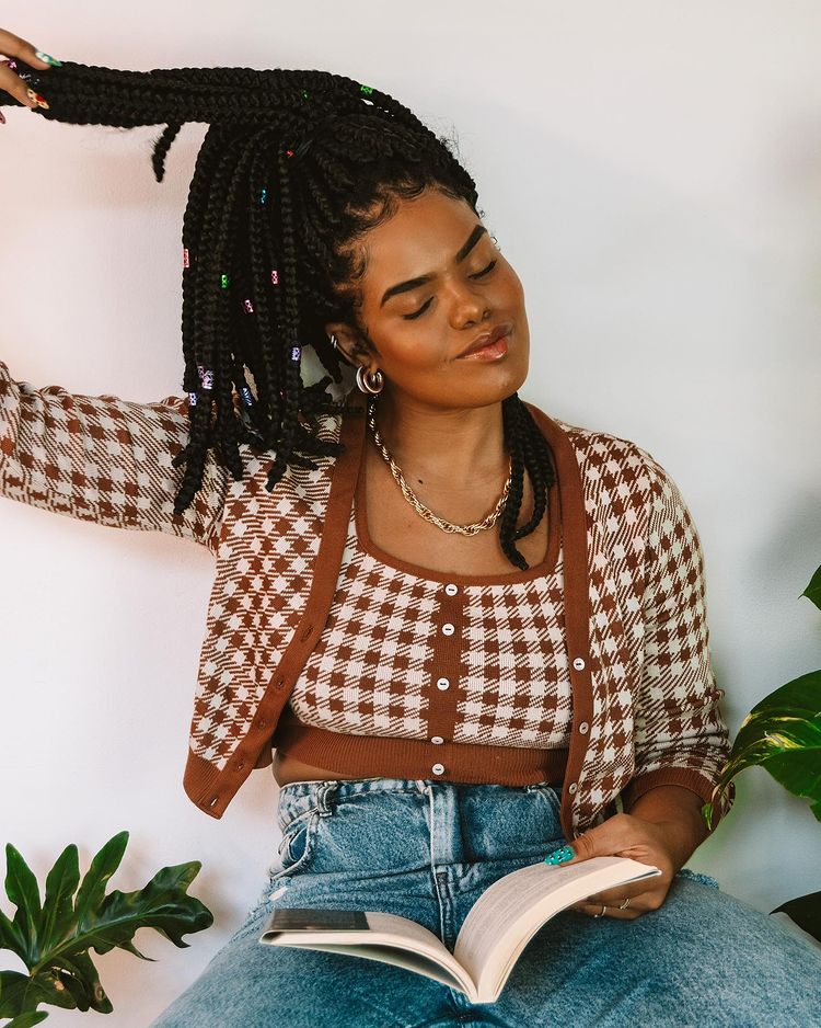 twin-set-cardigan-josyramos Garota usando top cropped com cardigan na mesma estampa xadrez marrom e calça jeans. Ela está sentada, com uma das mãos segurando um livro, e a outra no cabelo. Ela está de olhos fechados com a cabeça levemente inclinada para o lado.