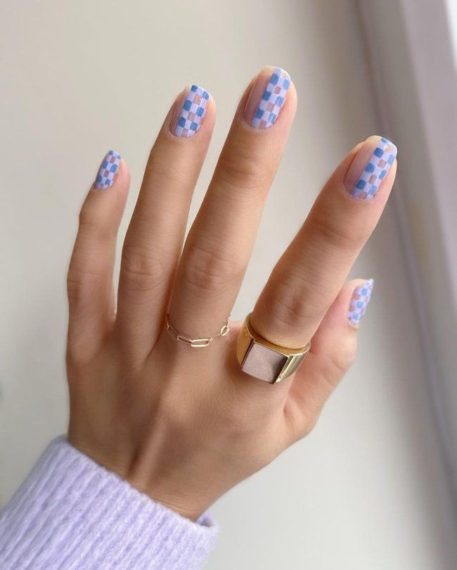 Foto de uma mão com anéis no dedo médio e indicador. A unha está com uma nail art xadrez azul.